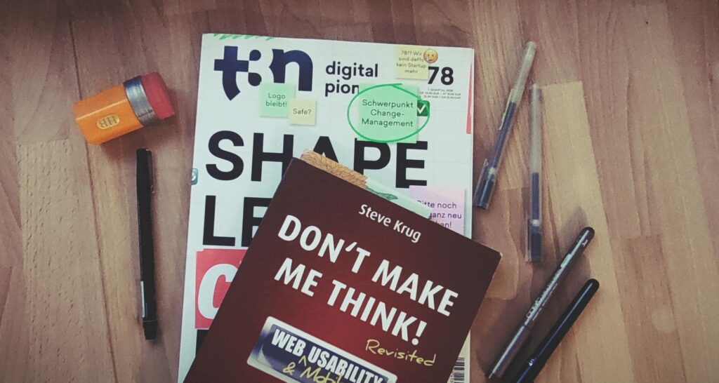 Eine Zeitschrift und ein Buch liegen auf einem Holztisch. Die Zeitschrift liegt unten. Es ist das t2n Magazin Nummer 78 mit dem Titel Shape, Lead, Change, Repeat. Darauf liegt das Buch "Don't make me think!" von Steve Krug. Um die Zeitschrift und das Buch liegen verschiedene Stifte und ein Spitzer in Form eines gelben Bleistifts.